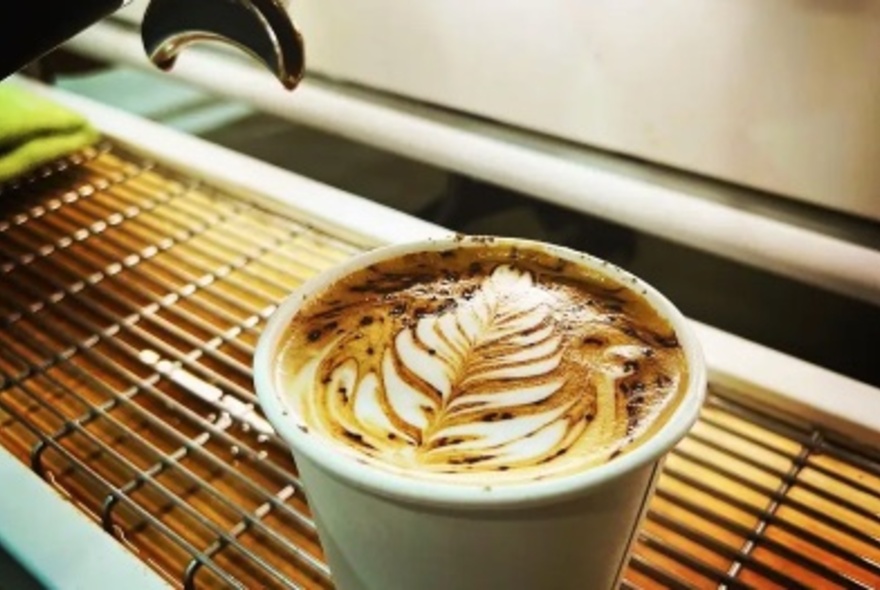A takeaway cup of coffee with milk froth art on the top, resting on the drain board of a coffee machine.