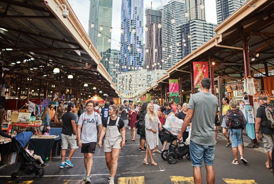 People walking through Queen Vic Market stalls with city buildings in the background.