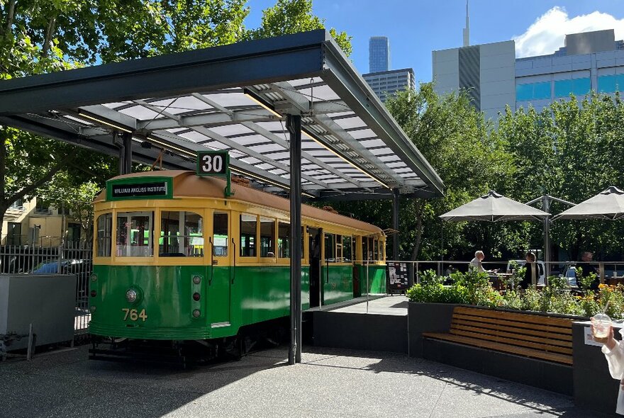 A yellow and green retro Melbourne tram parked under a roof with people sitting outside under umbrellas.