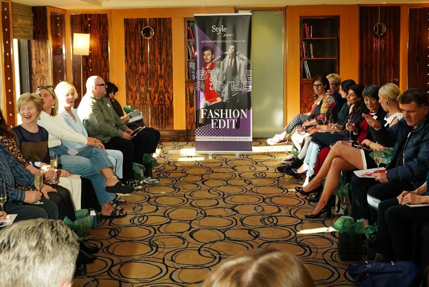 People sitting in a small room with patterned carpet with a sign saying 'Fashion edit' at the front. 