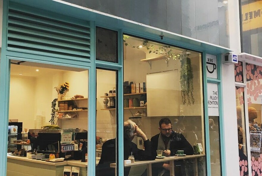 Small cafe with aqua painted windows and doorframes; two people sitting at window counter with coffee machine and counter behind.