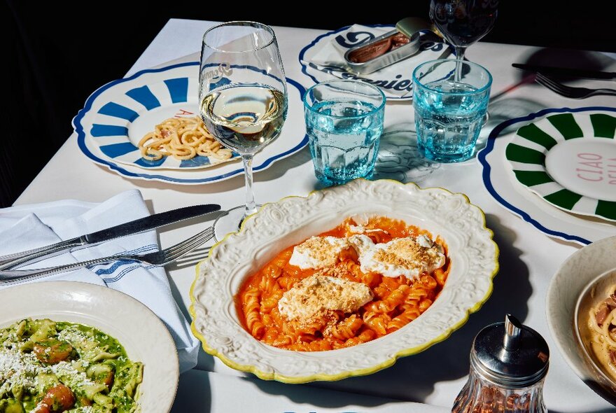 A share plate of tomato-based pasta surrounded by empty plates, wine and water glasses and cutlery on a white table.