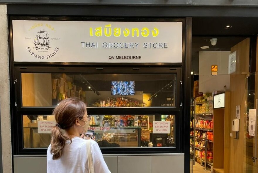 The exterior of Thai Grocery Store with signage, products in the window and a person standing outside the shop.