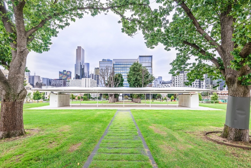 MPavilion, an open-concept building with a flat roof structure surrounded by grass and trees in front of the Melbourne cityscape.