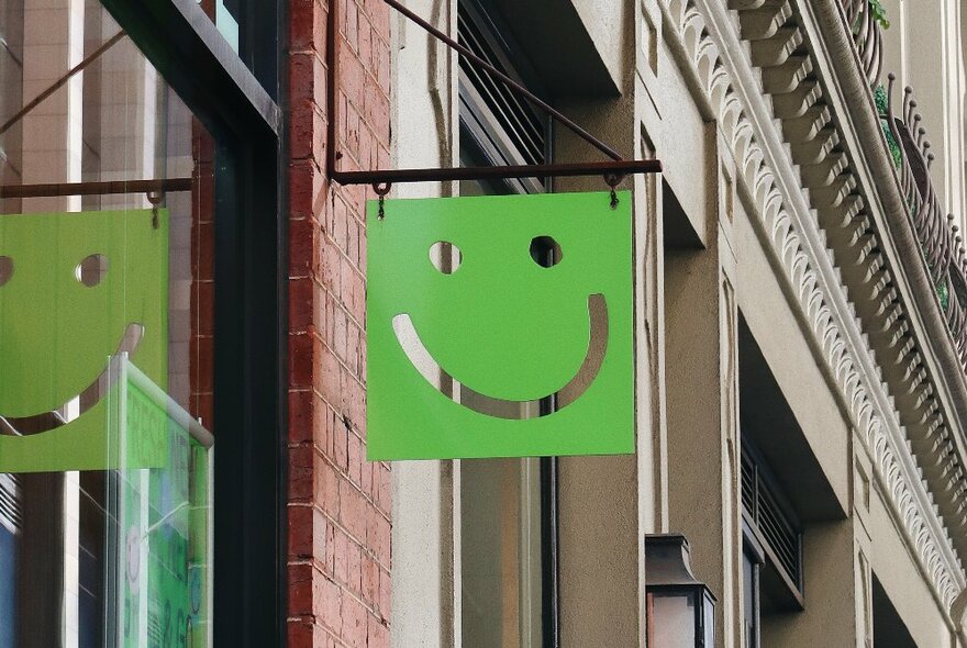 Green hanging sign with a smiling face, suspended from the brick wall of a heritage building.