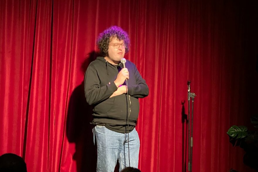 Comedian standing with a microphone on stage in front of red curtains.