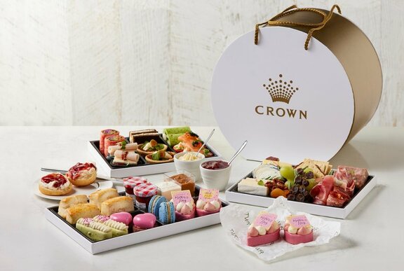 A round cardboard hamper box with the Crown Casino logo on it, behind a selection of high tea sweet and savoury treats.