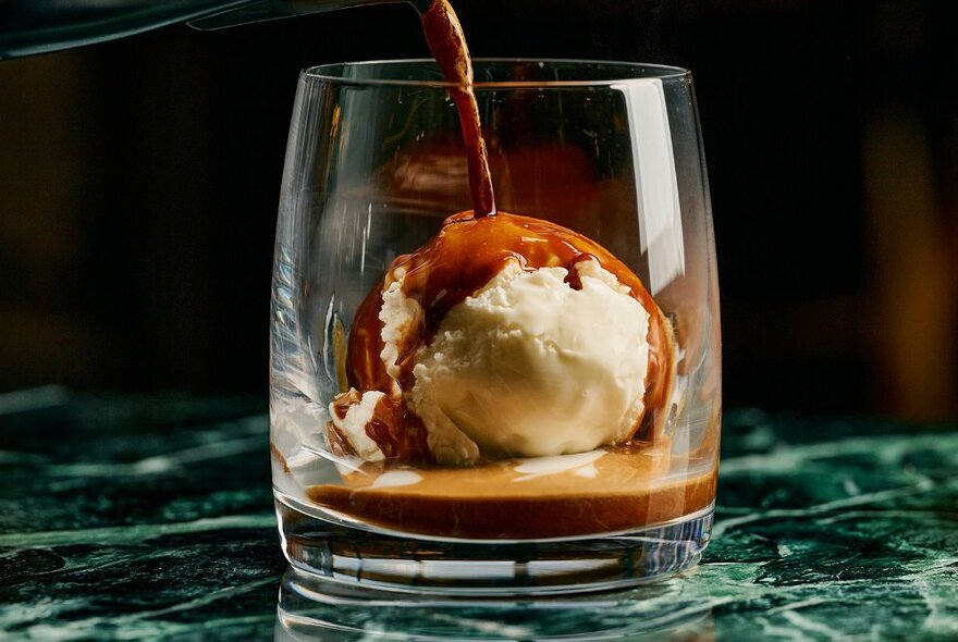 A lowball glass with a scoop of vanilla gelato and dark coffee being poured over the top to make an affogato coffee, on marble-green surface.