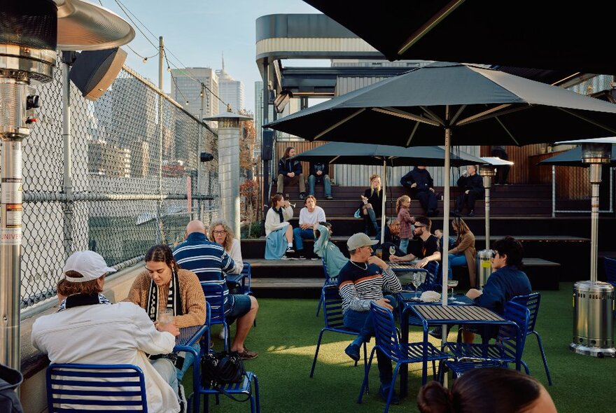 People enjoying drinks at a rooftop bar with green Astroturf, small tables and shade umbrellas, city buildings in the background.