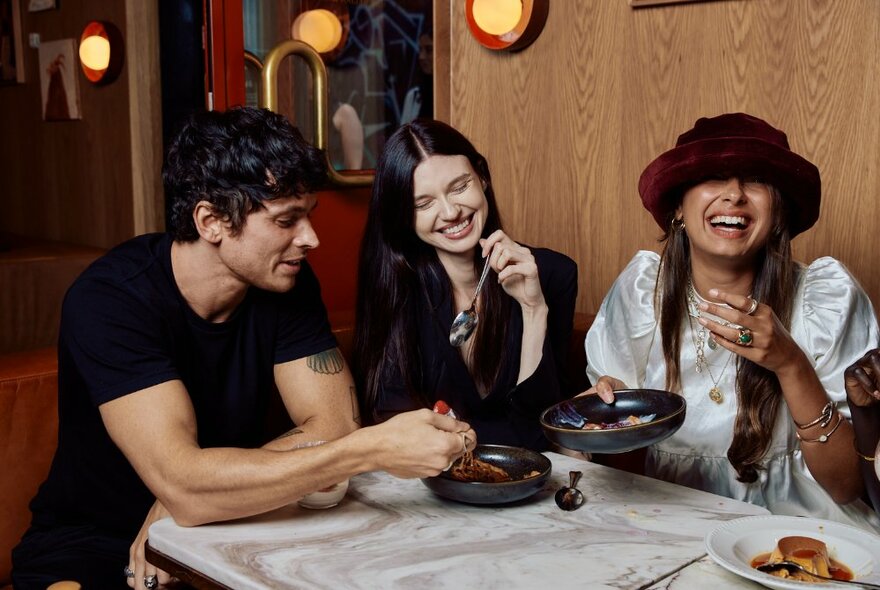 Three friends sitting at a table in a restaurant smiling and eating food from shared plates.