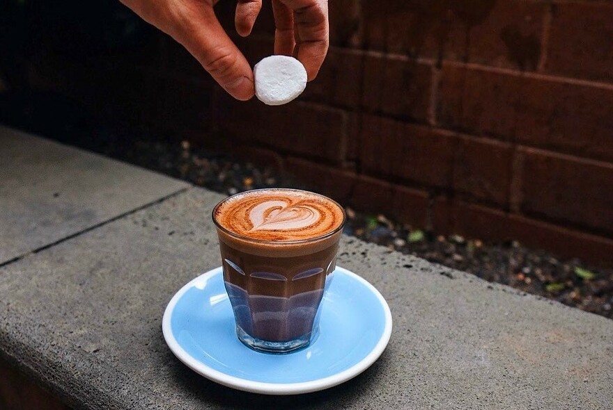 A hand dropping a marshmallow into a glass of coffee on a blue saucer.