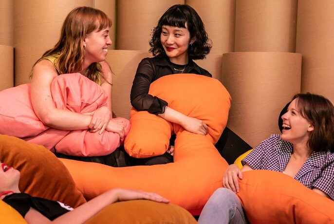 Four young adults smiling and interacting while sitting on bean bags and clutching orange and pink cushions.