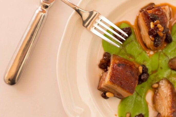 A segment of a plate containing cubes of pork belly and silver cutlery.