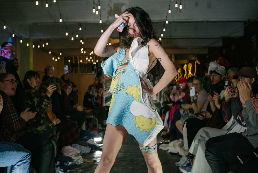 A person modelling clothes assembled from recycled fabrics, while onlookers clap hands and watch from seats either side of the runway.