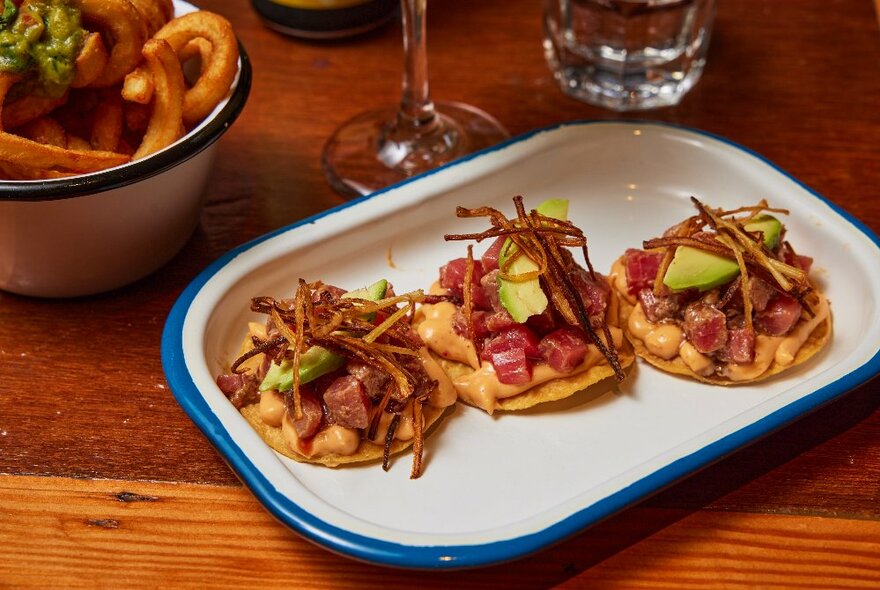 Three small tuna ceviche tacos on a white ceramic plate with a blue edge, on a wooden table with other plates and glassware behind.