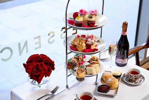 A three-tiered serving tray with a selection of treats, on a white table alongside a vase of red roses, a champagne bottle and other baked delicacies.
