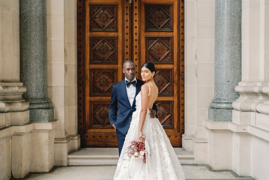 Bride and groom in wedding attire, posing for a photograph in front of a heavy carved oak door, framed by columns.