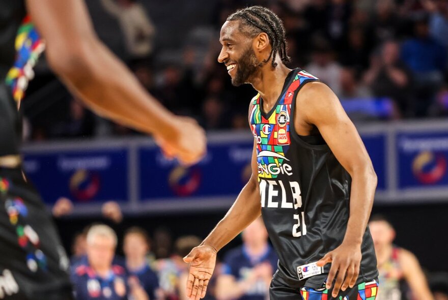 A Melbourne United basketball player smiling on the court, with out of focus players and fans in the background.