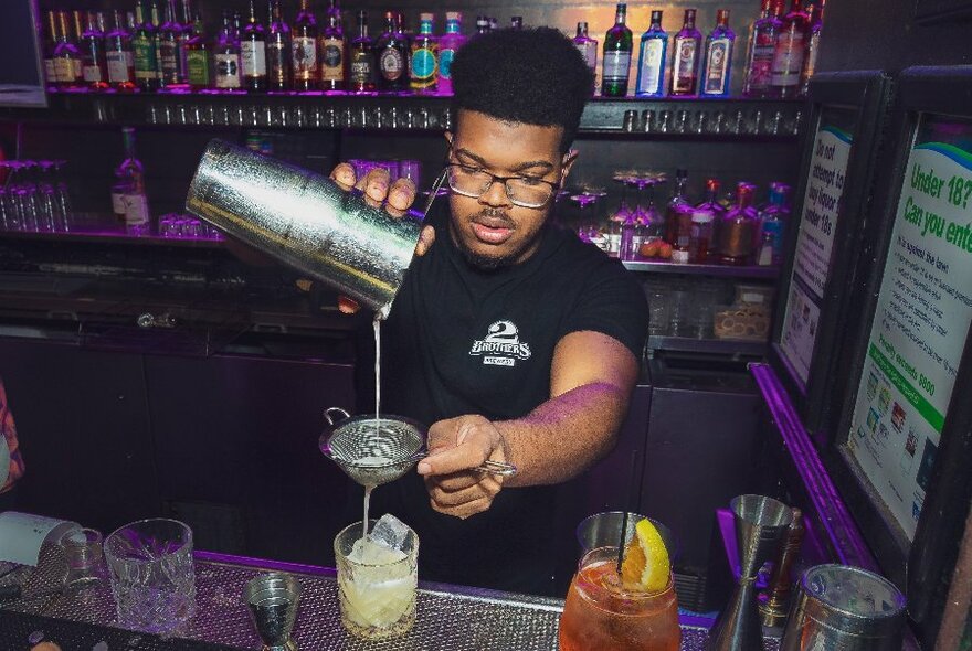Bartender pouring a cocktail from a metal shaker through a strainer.