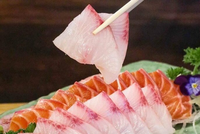 Chopstick holding thin slice of fish over platter of two types of fish.