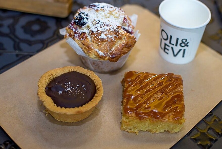 Three small food items including a muffin and a chocolate tart with a takeaway coffee cup resting on a counter.