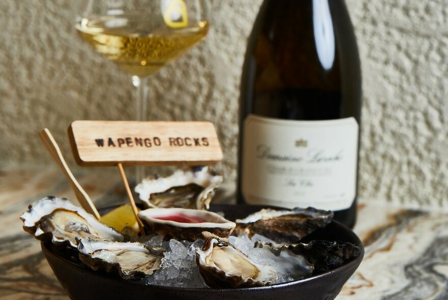 A bowl of oysters with a bottle of wine and a glass in the background.
