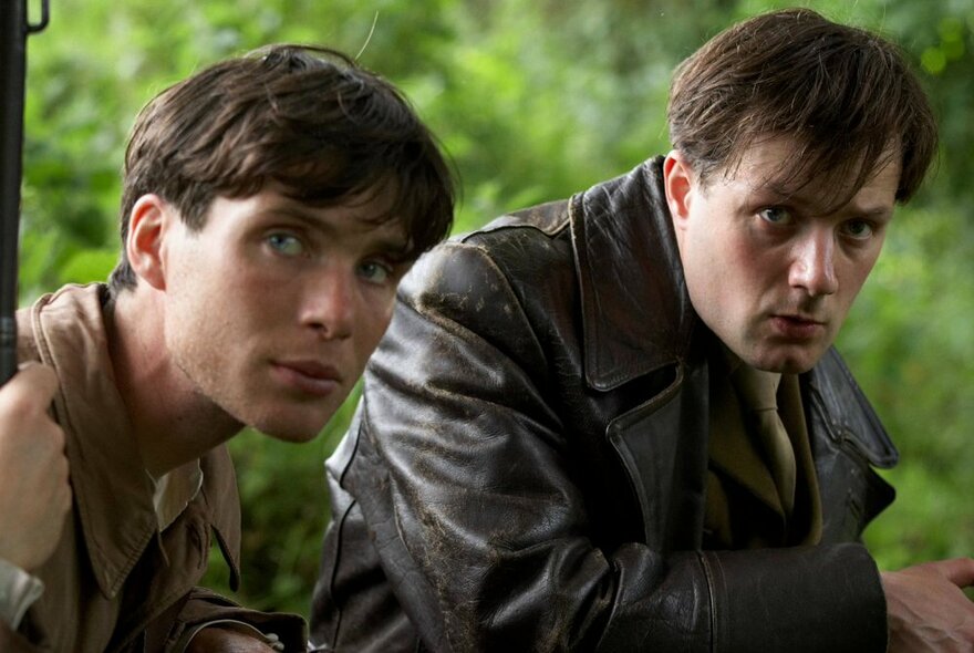 A still from a Ken Loach film featuring two scared-looking men crouching in a green field and looking at the camera. 