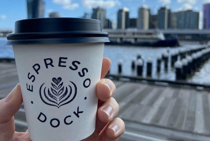 A hand holding a takeaway coffee cup up in front of a wharf and city skyline.