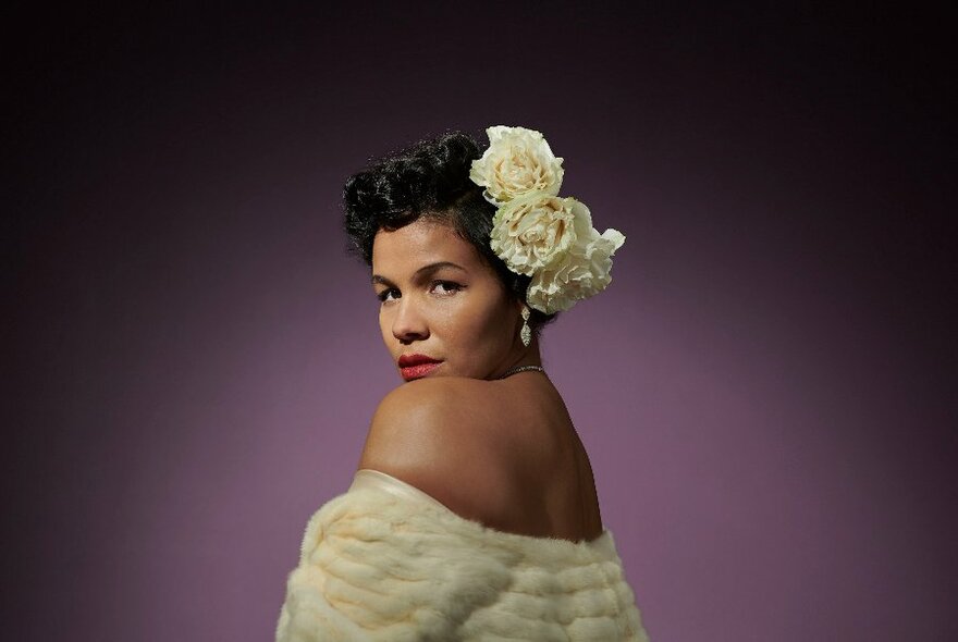Singer and performer Zahra Newman looking at the camera side-on, wearing a fur stole around her bare shoulders, and white flowers in her dark hair; posed against a purple backdrop.