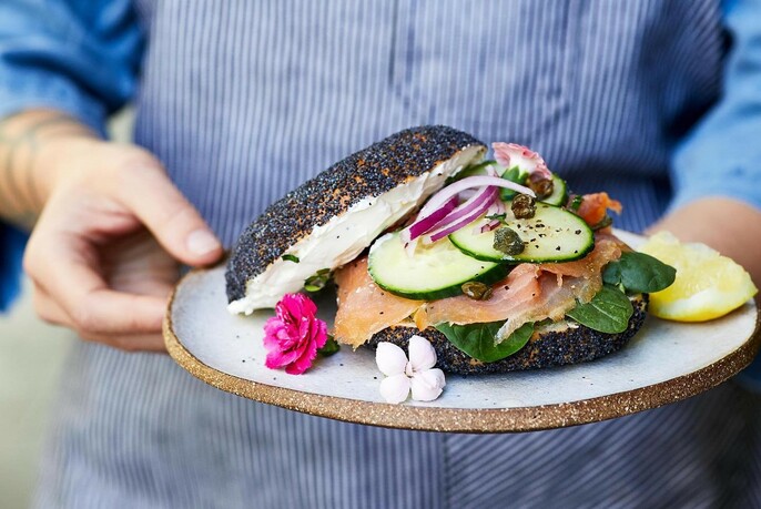 Waiter holding a rustic plate with poppyseed salmon sandwich garnished with flowers.