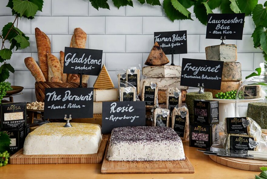 Large segments of assorted cheese displayed on wooden platters with hand written signs, on a counter, while in the background is a basket of breadsticks and greenery trailing along a white tile wall.