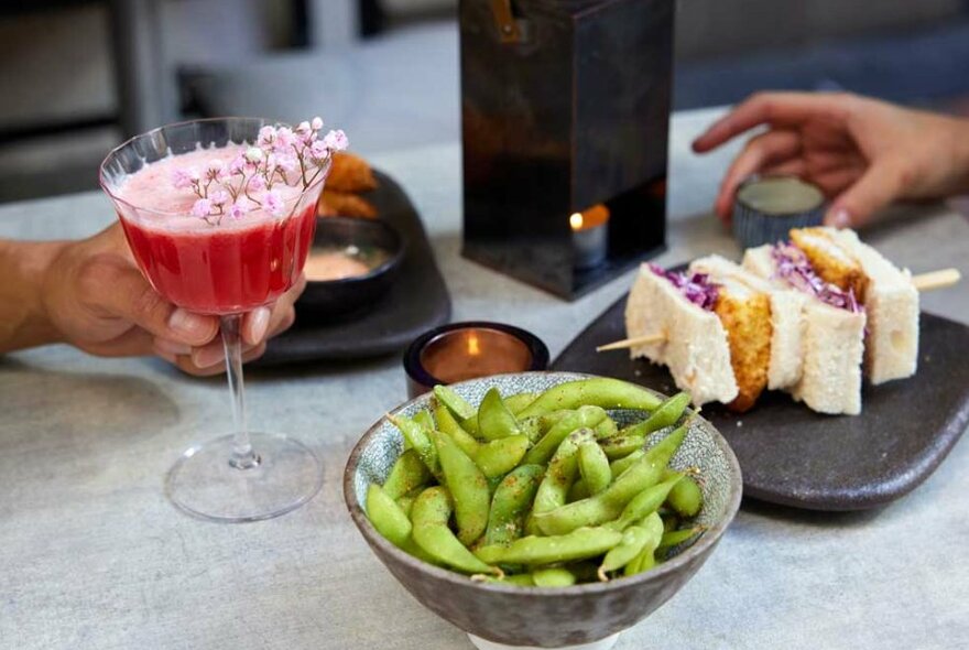 A pink cocktail next to a fried fish sandwich and a bowl of edamame