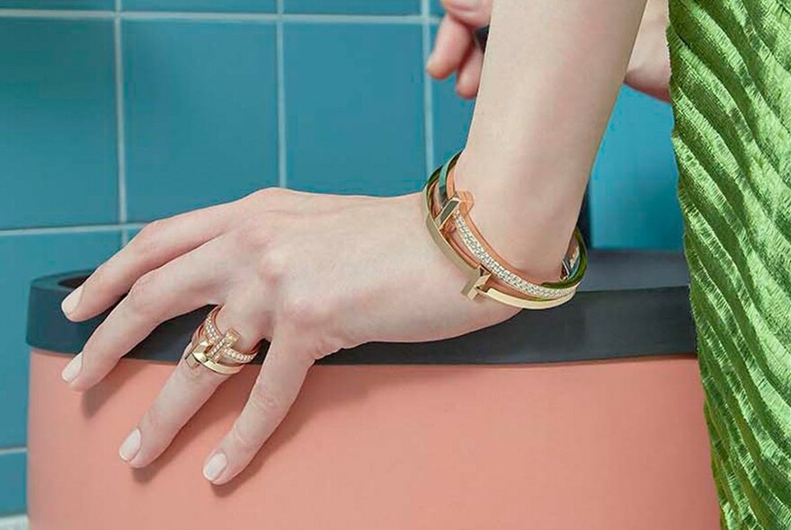 Model's hand with geometric gold and diamond rings and bracelets, with retro blue tiles in background.