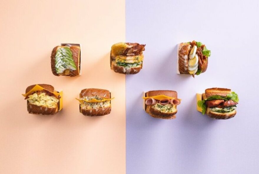Overhead view of seven toasted egg sandwiches with different extra fillings and garnishes on a pastel coloured background.