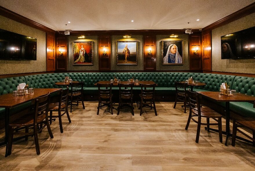 The interior of a traditional looking pub with a green upholstered curved banquette portraits on the wall. 