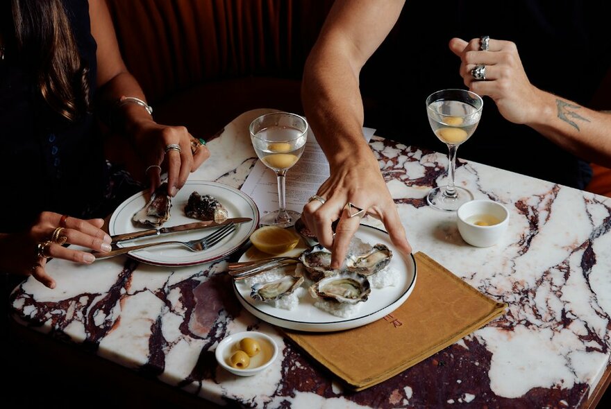Hands reaching in to select an oyster from a plate on a restaurant table, martinis also on the table.