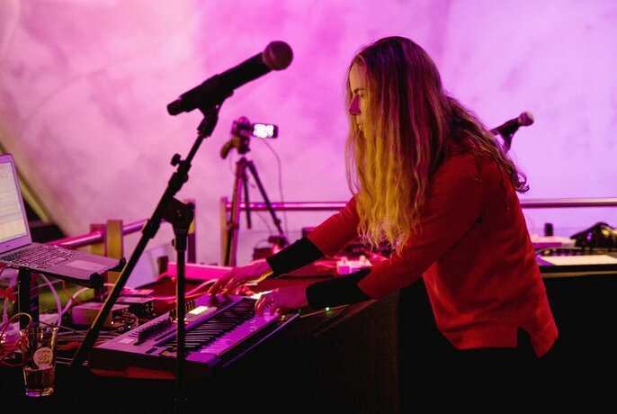 Person with long hair playing an electric keyboard, surrounded by audio equipment and a microphone.