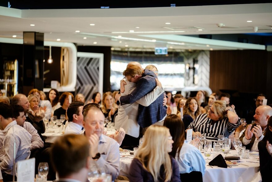 Two people embraced in a hug in front of a room of people seated at round dining tables who are watching and applauding.