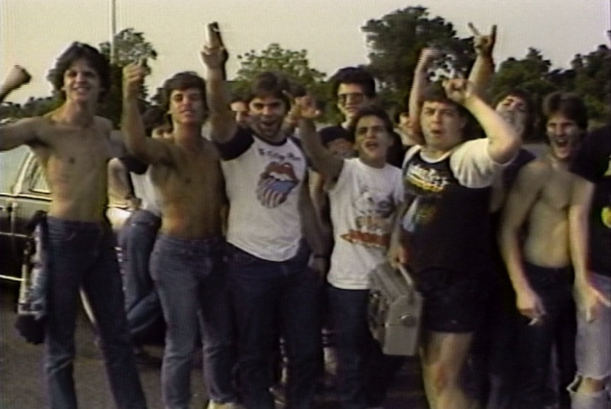 A still from a video of a group of young men in their late teens or early twenties, with their arms raised as if cheering, one holding a ghettoblaster or portable radio; standing in a car park.