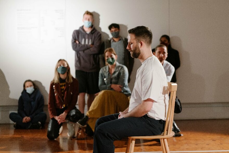A man sitting on a chair in the middle of a room while people wearing face masks look on from the sidelines. 