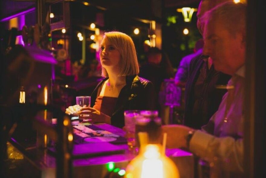 People standing with drinks at a bar under purple lights.