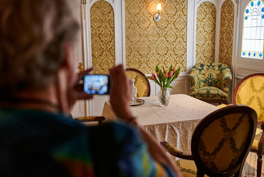 A woman in the foreground taking a picture of a full-size replica of one of the dining rooms on the Titanic, with ornate patterned wallpaper, a square table with tablecloth and flowers and three ornate chairs.