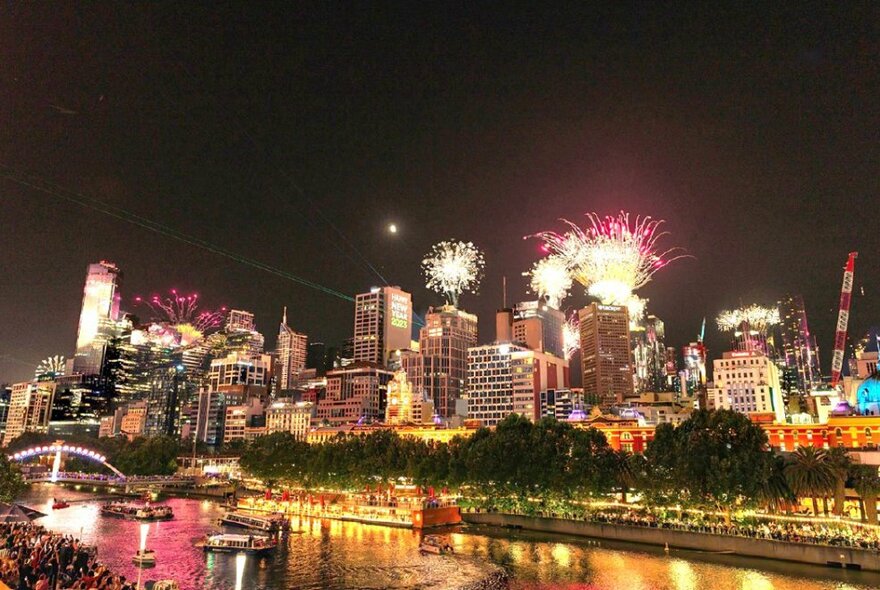 The city of Melbourne skyline at night, fireworks in the dark sky, the Yarra River reflecting the lights.