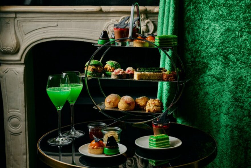 Black round table in front of fireplace with green cakes, sandwiches and green drinks on it.