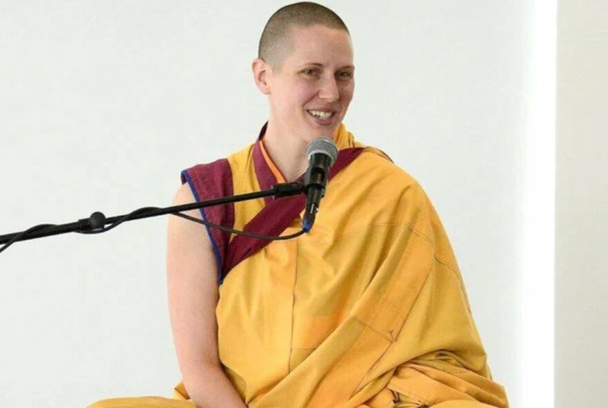 Buddhist woman wearing saffron robes, seated and speaking into a microphone.
