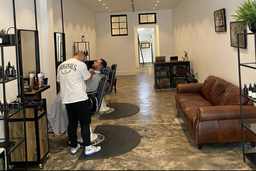 Interior of a barbershop showing a stylist trimming the beard of a client seated in a reclining chair, with couches, other chairs and mirrors on the wall.