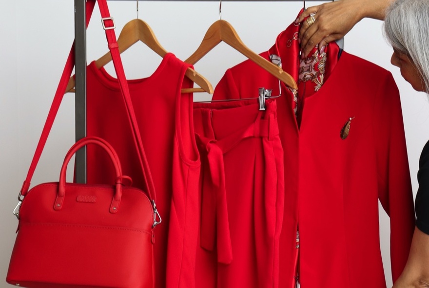 Display of red garments including a jacket, a skirt, a dress and a large red handbag hanging off a display rack, against a white background.