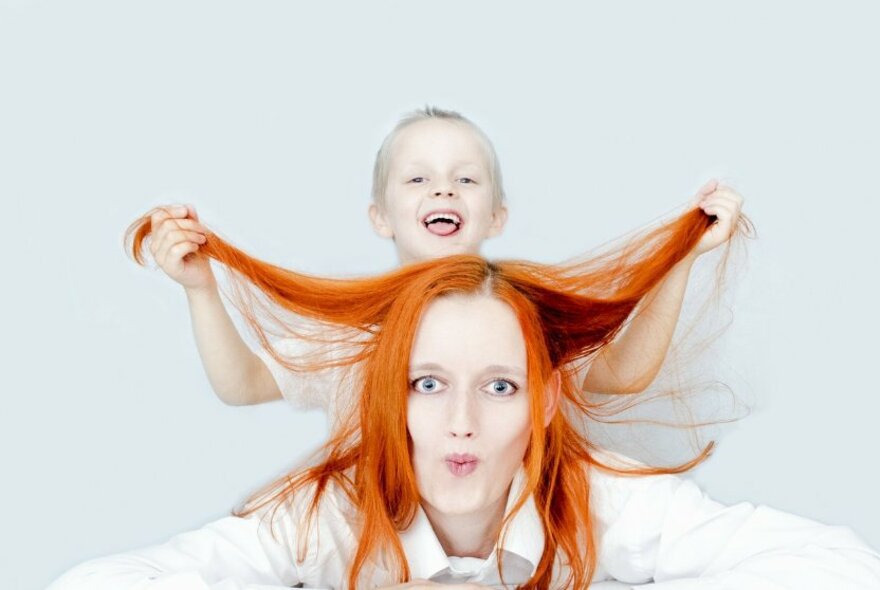 A woman with vivid orange hair being held up by a laughing todder behind her. 