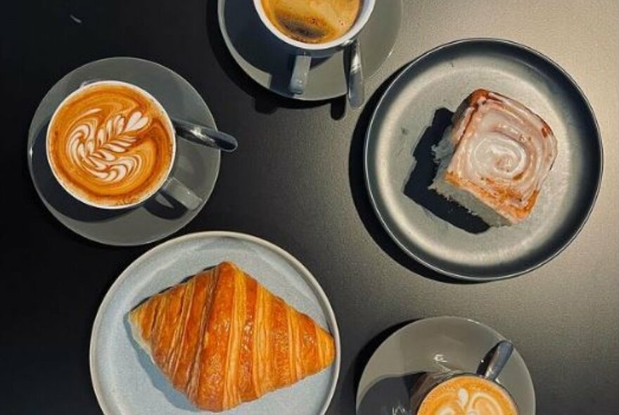 A croissant, three different types of coffee and a cinnamon scroll on plates on a black surface seen from above.