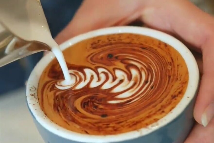 Milk being poured into a cup of coffee to create a pattern in the froth.
